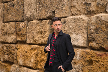 Portrait of young Spanish man, wearing black shirt, jacket and pants, with black handkerchief with red polka dots, posing next to a stone wall. Flamenco concept, art, dance, culture, tradition.