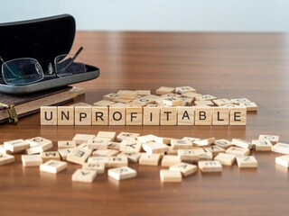 unprofitable word or concept represented by wooden letter tiles on a wooden table with glasses and a book - obrazy, fototapety, plakaty