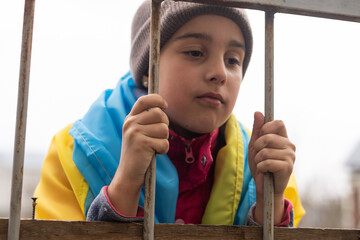 sad little girl with the flag of Ukraine behind a metal fence. Social problem of refugees and...