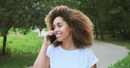 Cheerful biracial young female with curly hair talking use smartphone walking in park outdoor. Happy black skin woman laughing enjoying communication on mobile phone having positive emotion - Powered by Adobe