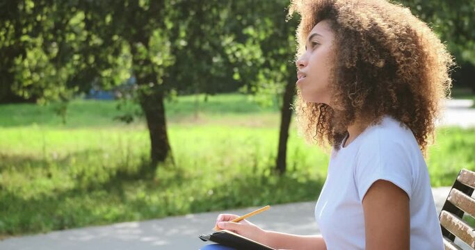 Pensive mixed race female handwriting paper notes in notepad page use pencil spending time alone sitting on bench at summer park. Thoughtful black skin young lady writing diary analyzing mental health