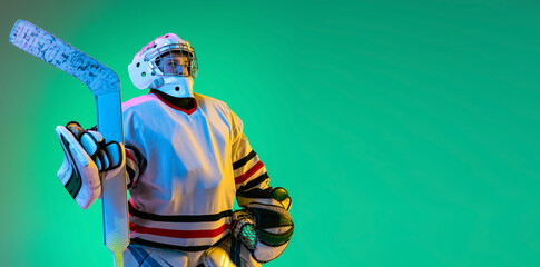 Portrait of boy, child, hockey player in uniform of goalkeeper posing isolated over green background in neon light