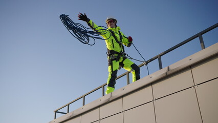 Industrial climber throws rope skein down from roof of building. Man prepares for climbing down skyscraper against cloudless sky low angle shot