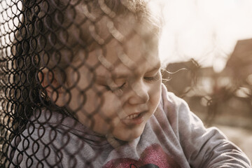 A little girl with a sad look behind a metal fence. The problem of forced deportation of Ukrainian...