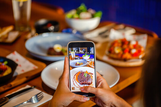 Woman photographing gourmet steak through mobile phone at luxury restaurant table. Hand hold smartphone and taking photo of meat on dish before eating in cafe.