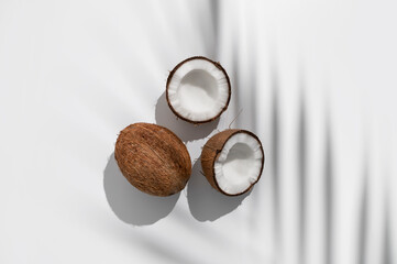 Fresh whole and broken pieces tropical coconut nut with sunny contrast shadows palm leaf on light gray background flat lay top view. Creative summer food background, exotic organic healthy diet fruit