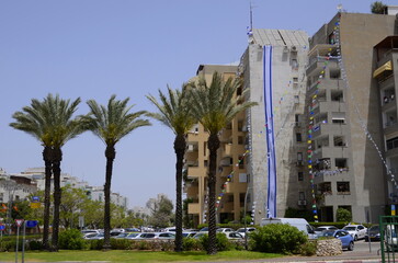 Israel Independence Day. The city is decorated with flags. An ordinary house and a huge flag of Israel. Palm trees next to a multi-storey building.