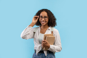 Portrait of young black female student with books touching glasses and smiling at camera over blue...