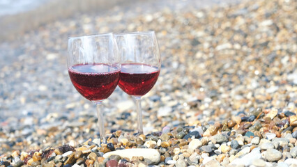 Two glasses of red wine on the pebble beach at the sea coast. Concept. Vacation pleasure and romantic date details, close up of two transparent glasses standing on stones near wavy shore.