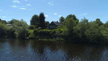 Obraz premium Aerial view of a river in summer village and an old wooden house. Shot. Forest and small house surrounded by green trees and bushes along the river bank on blue clear sky background.