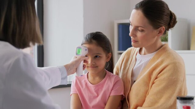 medicine, healthcare and pediatry concept - mother with sick little daughter and doctor measuring temperature with infrared forehead thermometer at clinic