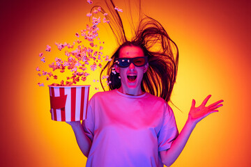 One excited young girl, student wearing 3d glasses and holding bucket of popcorn isolated on orange background in neon light, filter. Concept of emotions, youth, beauty