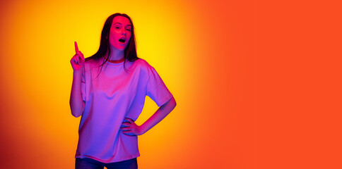 Flyer with portrait of young charming girl, student with long hair posing isolated on orange background in neon light, filter. Concept of emotions, youth, beauty