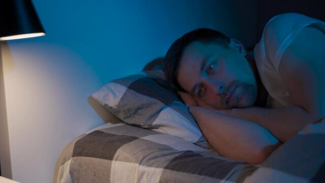 Caucasian man suffering from insomnia, lies in bed at night with open eyes and looks at one point. Topic of sleep disorders in emotional stress, fear and panic attacks. I can't sleep for long time.