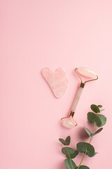 Beauty procedure concept. Top view vertical photo of eucalyptus gua sha and rose quartz roller on isolated pastel pink background with copyspace