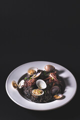 Black risotto with cuttlefish ink and seafood: clams and mini squid on a white plate, dark key, black background. Mediterranean, Italian seafood recipes, front view