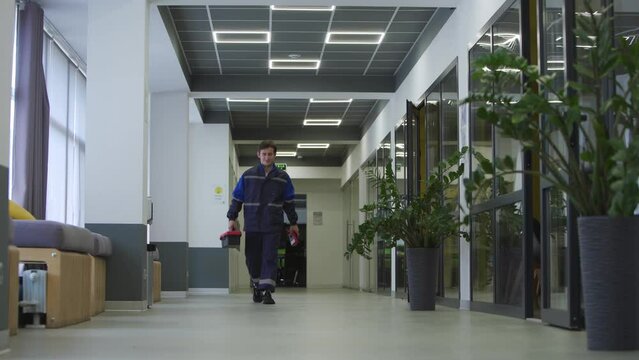Electrician repairs light in office. Master finishes work with electricity. Locksmith walking along business center hall with toolbox. Man goes to apply for job as engineer. Male mechanic in uniform.