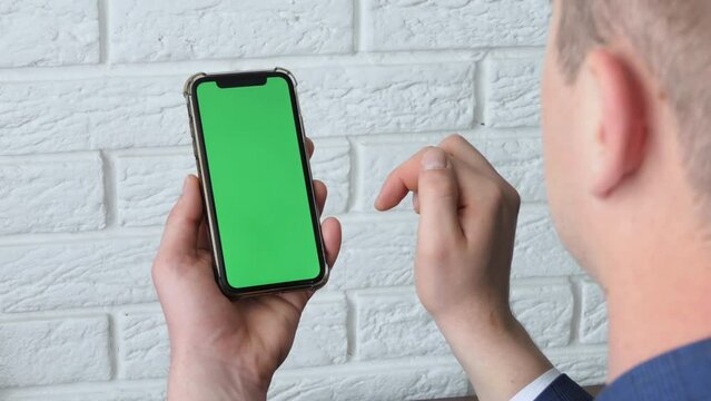 Man using smart phone in office space, sitting on chair and using green screen phone mockup, chroma key, browsing content, video, blogging, tapping center of screen, flipping through news flyer.