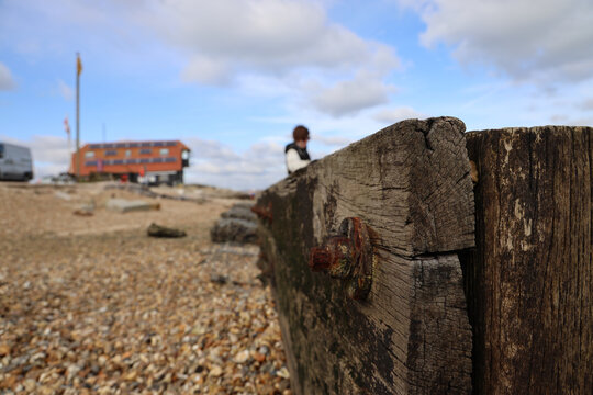 Wooden Sea-breakers On A Pebble Beach Stretching Into Distance, A Person Out Of Focus Leaning On End