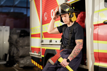 Photo of fireman wearing helmet with ax against fire engine. Image of young man firefighter with...