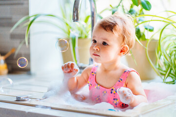 Cute baby girl with big eyes playing with foam and bursting soap bubbles in kitchen sink next to...