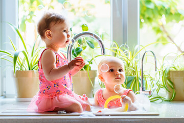 Two cute baby girls playing with foam and toys and bursting soap bubbles in kitchen sink next to...