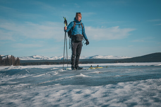 Young white man with skis and backpack and blue down jacket on frozen lake Storsjö with mountain view (onto Storådörren) close to Ljungdalen (Sweden).