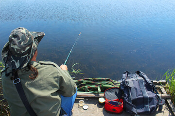A fisherman with a fishing rod sits on the river bank, rear view on a summer day. Catching fish from the lake on a sunny day
