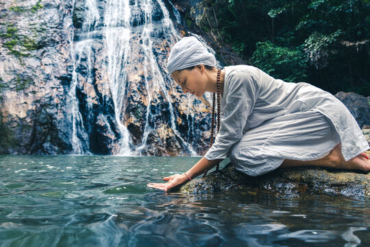 A young woman in white clothes and a turban sits on the rocks near the waterfall and scoops up water with her hands.