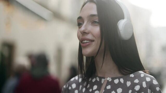 Attractive young woman listening to music in headphones standing in the city street. Portrait of beautiful brunette.
