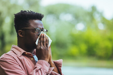 Allergic black man blowing on wipe in a park on spring season. Man with allergy or cold, blowing...
