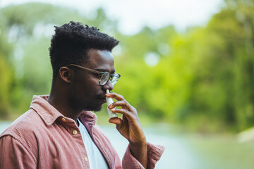 A Man is Walking in Public Park and Using a Nasal Spray Due to the Problems with Breathing or Allergy. Young African-American Man is Having Sinusitis Problems Outside in Nature.