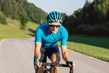 Caucasian man in professional road cycling clothes, a blue jersey, with a helmet, sunglasses, and gloves, riding uphill on an asphalt road