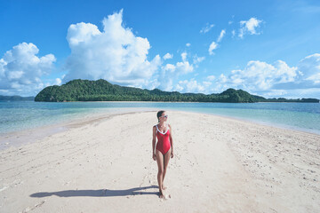 Fototapeta na wymiar Vacation on the seashore. Young woman in red swimsuit walking on the beautiful tropical white sand beach.