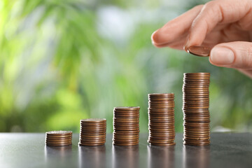 Woman stacking coins at grey table against blurred green background, closeup. Space for text