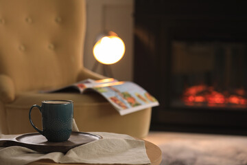 Cup of hot drink on table near fireplace at home, space for text. Cozy atmosphere