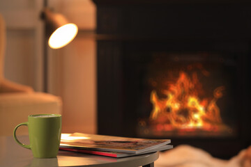 Cup of hot drink and magazines on table near fireplace at home, space for text. Cozy atmosphere