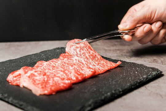 Premium Rare Slices sirloin Wagyu A5 beef with high-marbling texture pick up by bbq tongs with hand from stone plate. Served for Yakiniku, Sukiyaki and Shabu. Image with copy space.