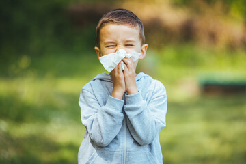 Little blond boy sneezing due to allergy related problems, on a sunny day outdoors. He is holding a handkerchief in his hands, looking away. Copy space available.