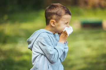 Child with pollen allergy. Boy sneezing and blowing nose because of seasonal allergy while sitting in a grass. Spring allergy concept. Flowering bushes and trees in background. Child allergy - obrazy, fototapety, plakaty