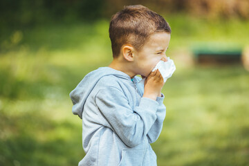 Fototapeta premium Little blond boy sneezing due to allergy related problems, on a sunny day outdoors. He is holding a handkerchief in his hands, looking away. Copy space available.