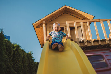 Fototapeta na wymiar Happy little boy play outdoor on the children playground and ride the yellow plastic children's slide in the summer 
