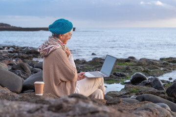 Smiling caucasian senior woman in remote work sitting at the beach using laptop. Elderly white haired lady enjoying sea vacation video calling with computer