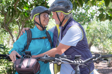 Fototapeta premium Happy senior caucasian couple hug in public park while cycling in nature with their e bike. Lovely elderly couple looking into each other eyes