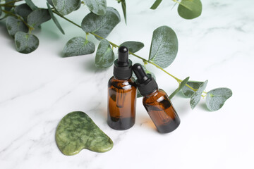 Glass bottles with oil, jade Gua sha stone for face massage on marble background with eucalyptus...