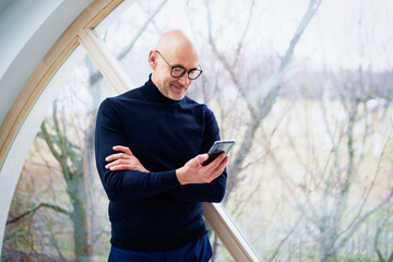 Confident man using mobile phone and text messaging while standing by the window