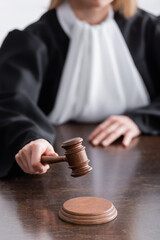 partial view of judge in black mantle holding wooden gavel on blurred background.