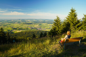 A woman sitting on a bench, looking from mount Hohenbogen to Neukirchen Heiligblut, a small town in...
