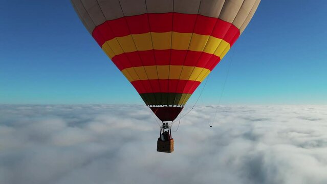 Closeup colorful red yellow hot air balloon with happy tourists flying above white clouds. First person adventure view from drone.
