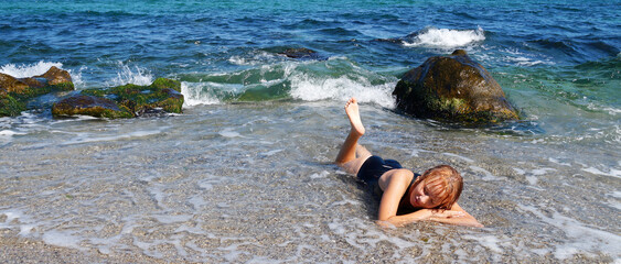 a teenage girl in a black bathing suit is sunbathing on the shore lying in the water